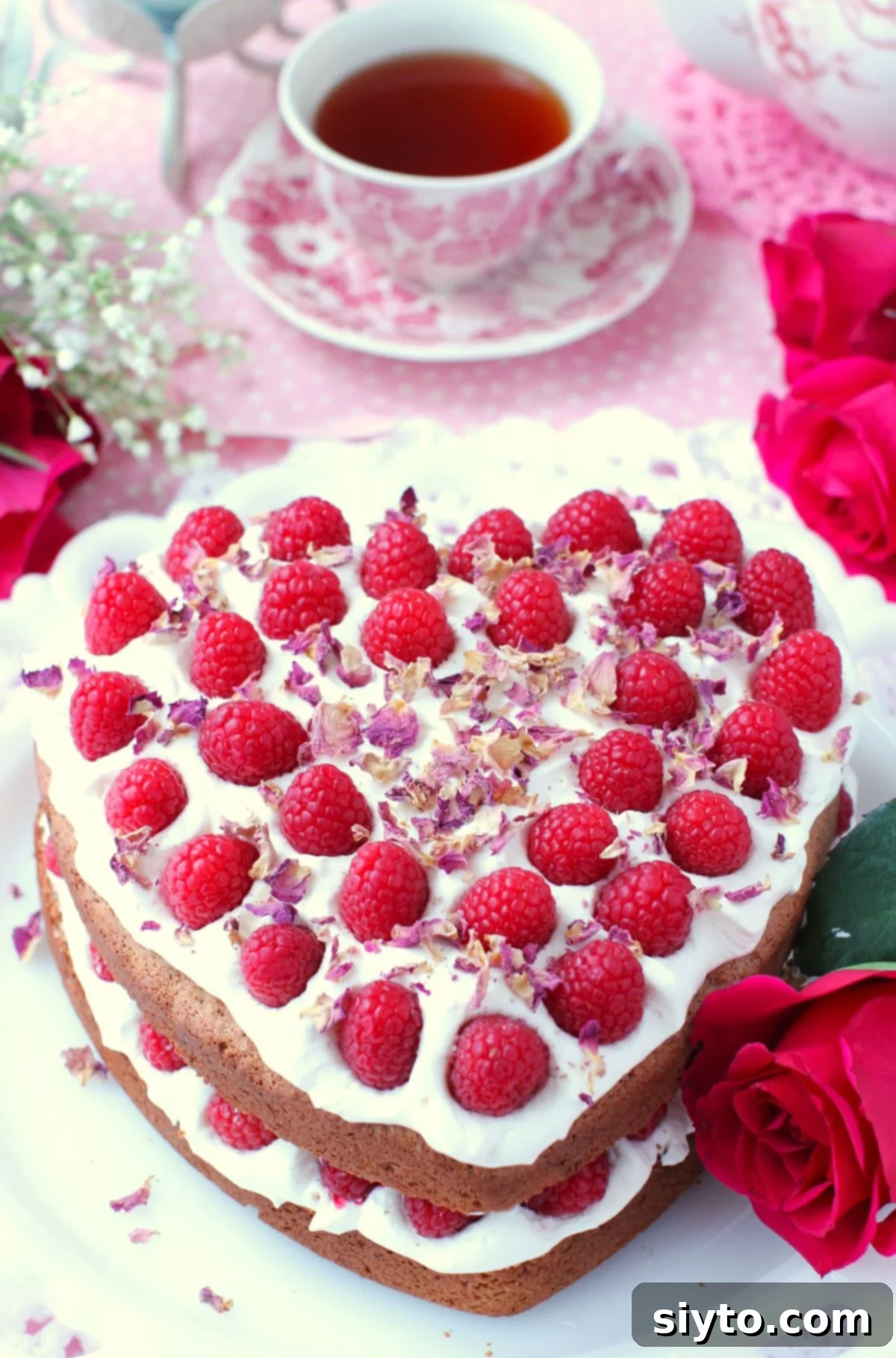 The full Raspberry Rose Heart Cake, beautifully adorned with fresh raspberries and rose petals, resting on a white cake stand with a cozy cup of tea in the background, signaling a moment of sweet indulgence.