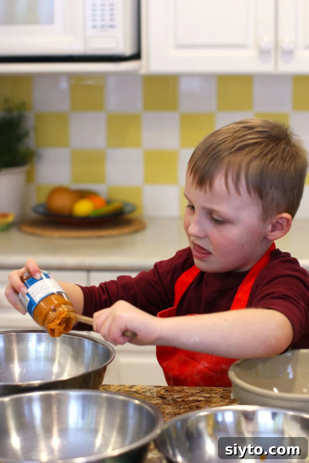 Caleb, a young boy with focused expression, carefully scooping baby food out of a glass jar into a stainless steel mixing bowl.