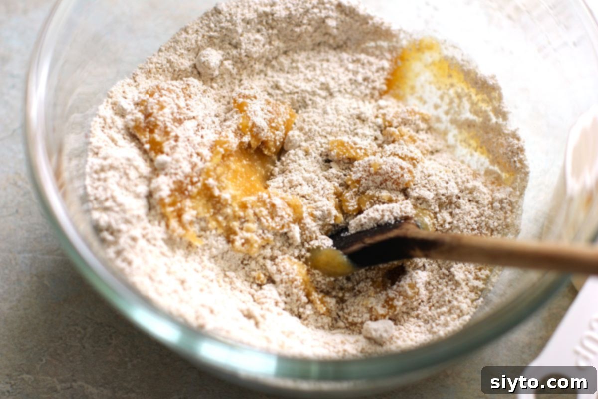 A close-up shot of flour and baby food being mixed in a clear glass bowl with a wooden spoon, showing the beginning stages of dough formation.