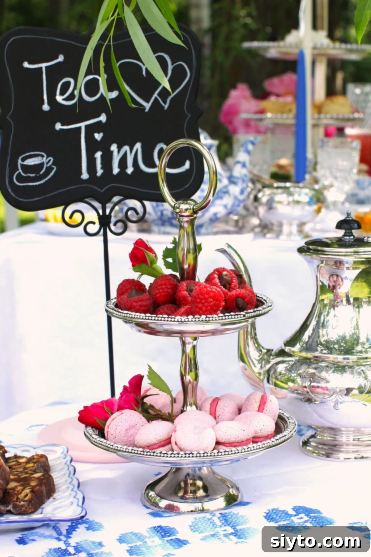 Silver two-tiered cake stand with Raspberry Meringue Kisses on the bottom and ganache-filled raspberries on the top, silver teapot besided and Tea Time sign behind.