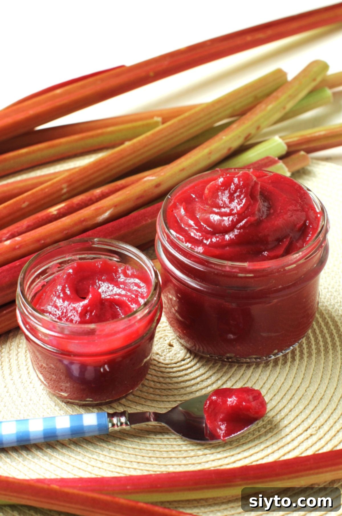 Two jars of homemade rhubarb butter, one large, one small, with the rich, ruby-red spread swirled high. Fresh rhubarb stalks are artfully arranged behind the jars, and a spoon filled with the delectable rhubarb butter sits invitingly in front.