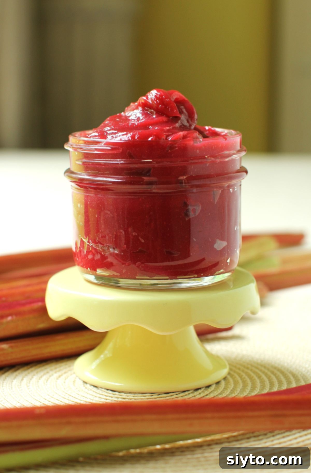 A jar overflowing with a mound of rich, ruby-red rhubarb butter, elegantly placed on a small footed yellow stand. Fresh rhubarb stalks surround the base, enhancing the natural beauty of the scene.