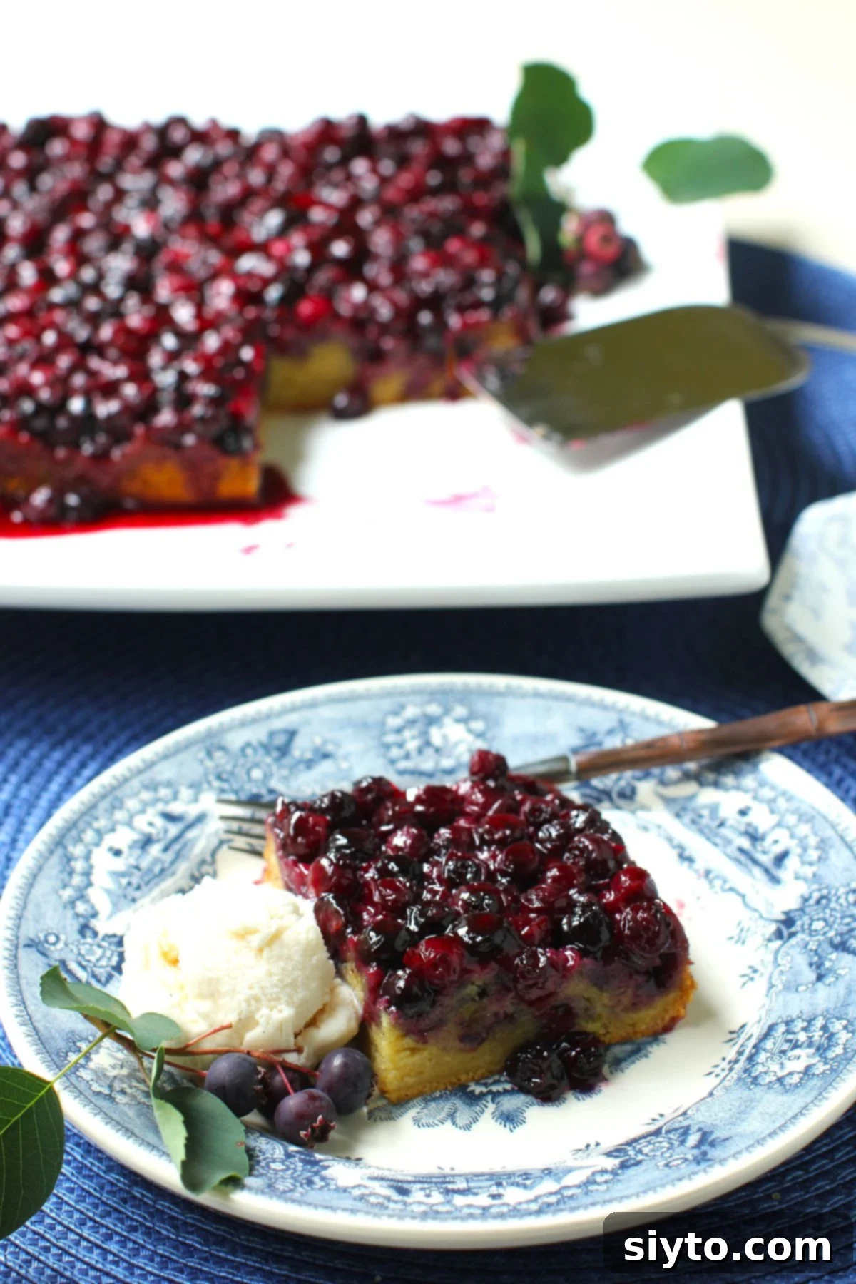 Square of Saskatoon Upside Down Cake on a blue and white plate, with a scoop of ice cream beside it and the square platter with the whole cake behind it.