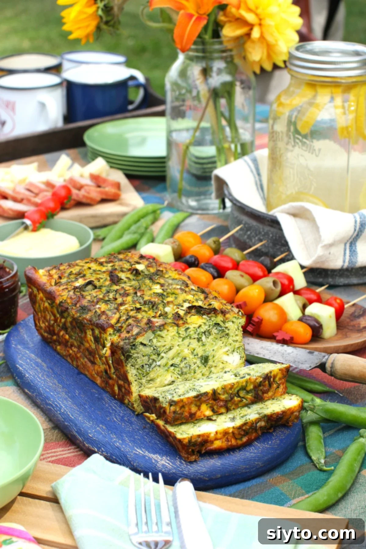 A golden-brown baked zucchini frittata loaf, partially sliced and resting on a blue cutting board, surrounded by an array of colorful picnic items, including fresh fruits and vegetables.