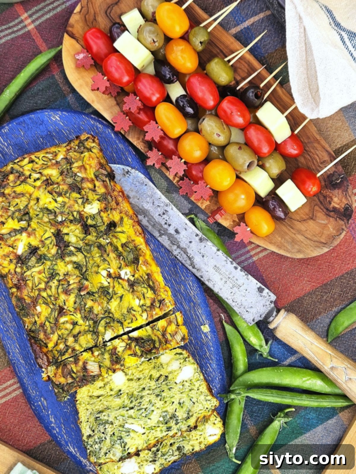 A Zucchini Frittata Loaf artfully arranged on a blue board, alongside a wooden platter showcasing vibrant cherry tomato, cucumber, and olive skewers.