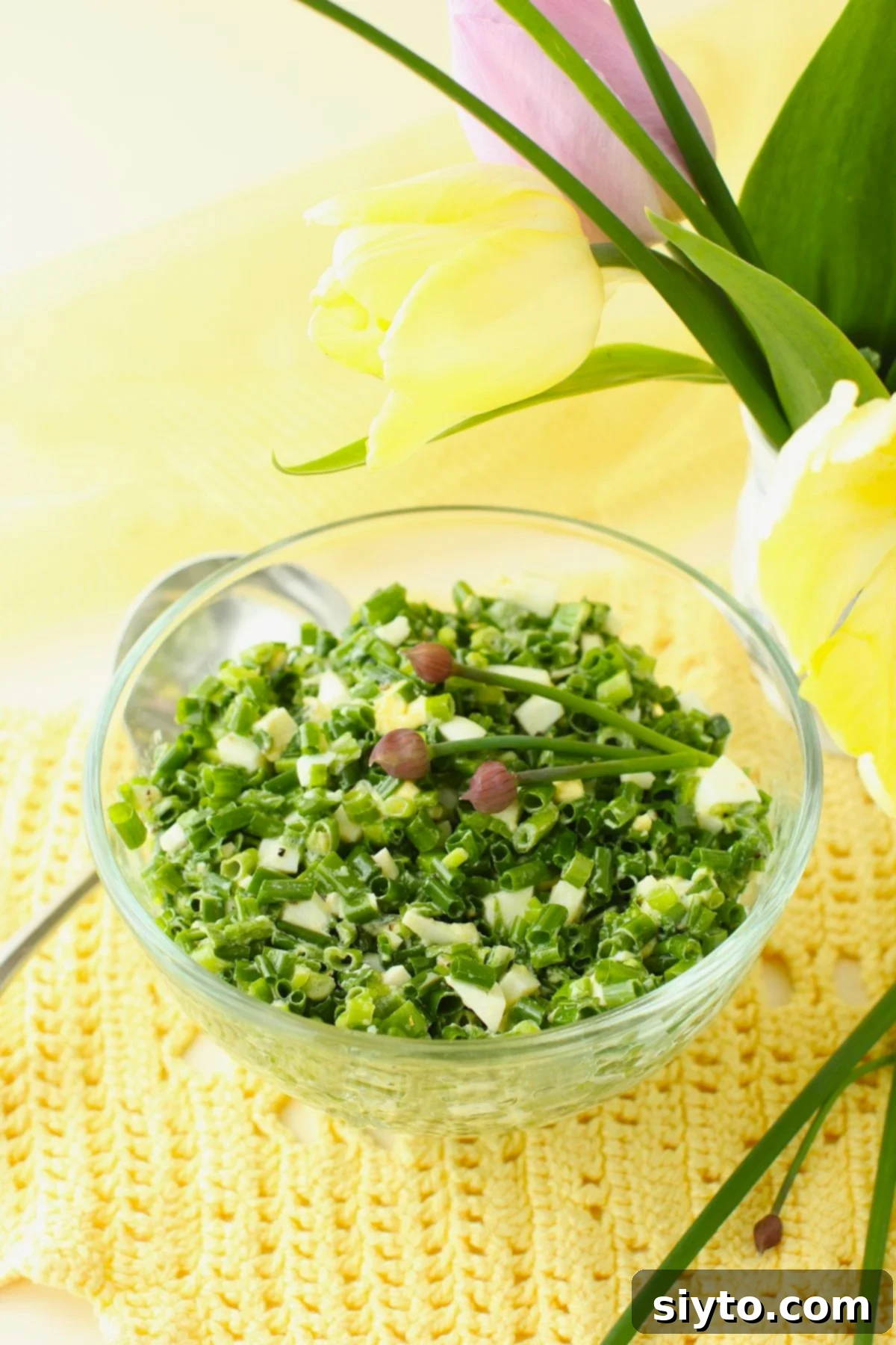 Clear glass bowl of German Chive Salad (Schnittlauchsalat), garnished with chive buds, resting on a charming yellow crocheted doily.