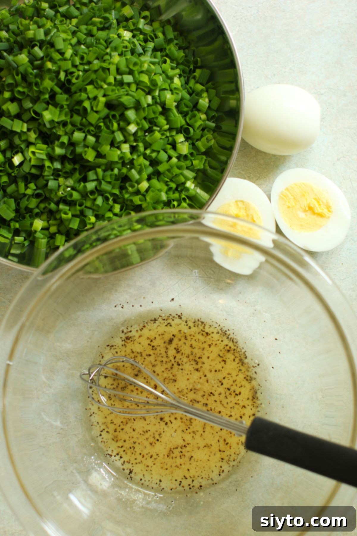 Bowl of finely chopped fresh chives and two hard-boiled eggs beside it, with dressing ingredients whisked in a separate small bowl.
