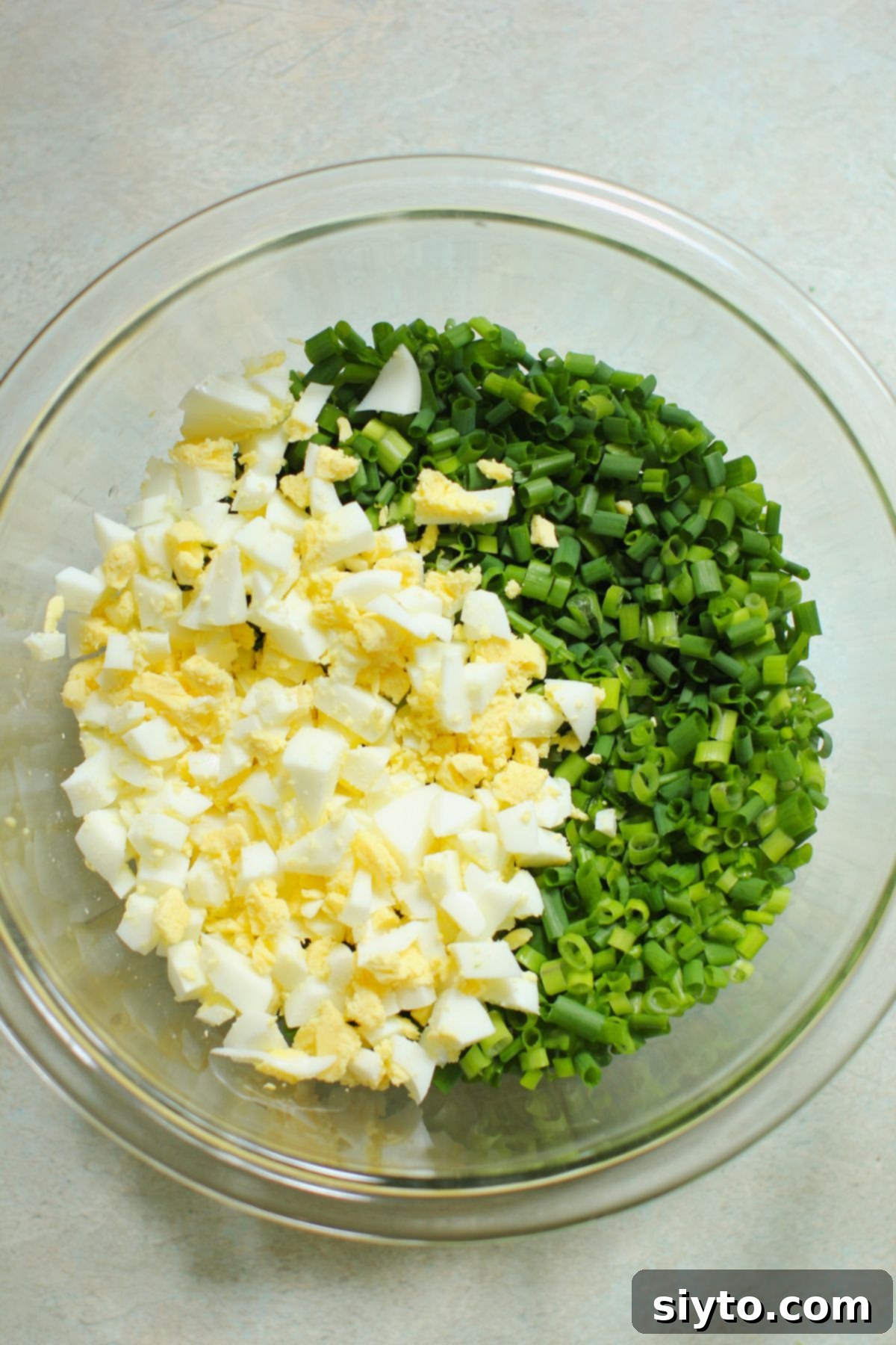 Chopped chives and hard-boiled eggs piled on top of the whisked dressing in a clear bowl, ready to be mixed.