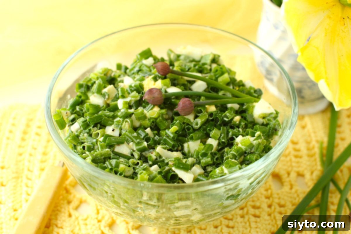 Close-up of a clear glass bowl of German Chive Salad (Schnittlauchsalat) garnished with delicate chive buds, against a soft yellow background.