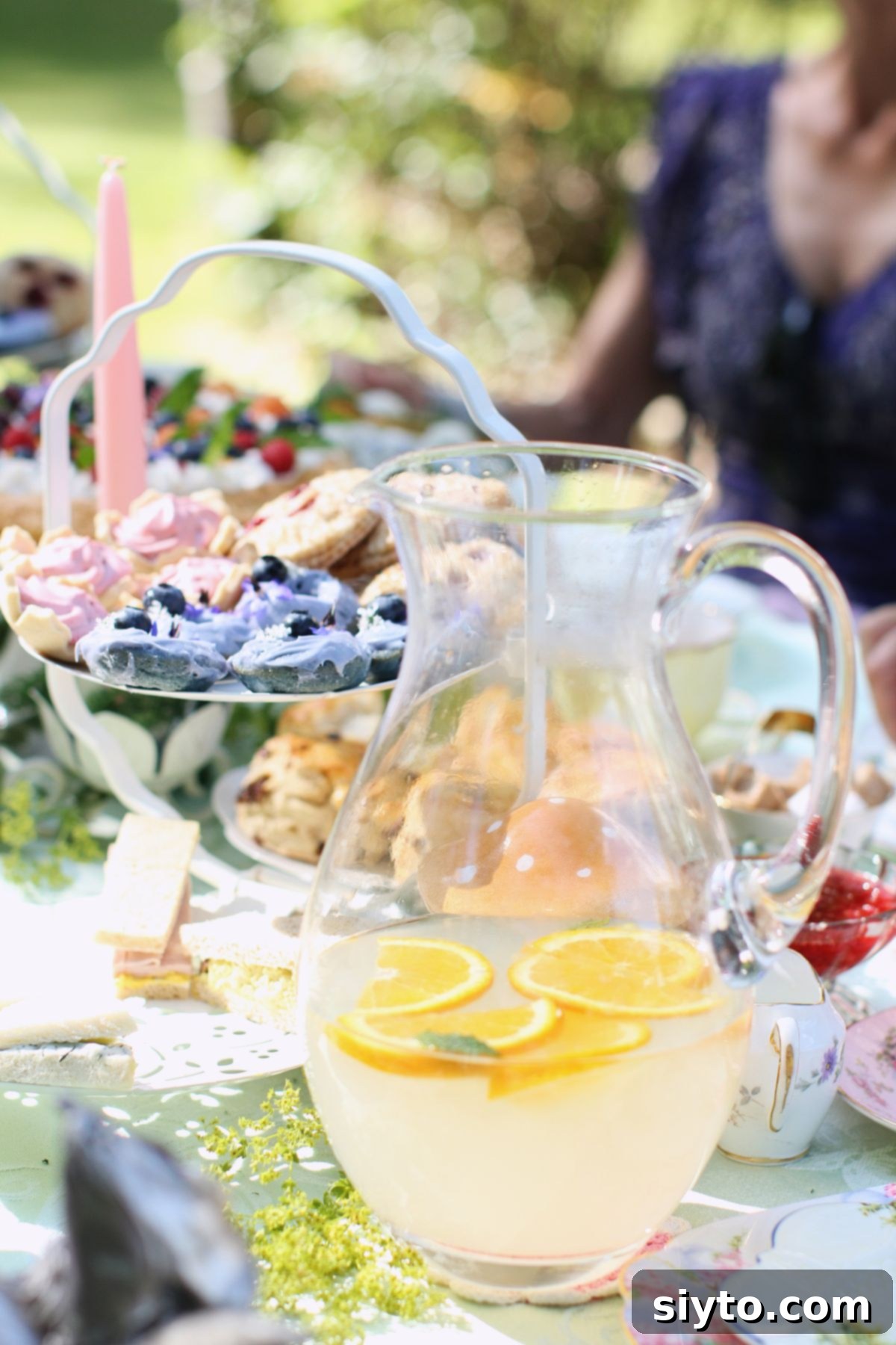 A beautifully set tea table laden with various goodies, including mini blue donuts prominently displayed on a three-tiered cake stand. A pitcher of refreshing lemonade sits invitingly in the foreground.