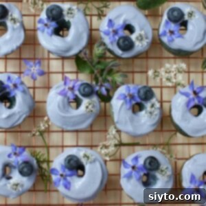 Square thumbnail photo. Mini blue donuts laid out in a grid pattern on a copper cooling rack, with borage flowers and cilantro flowers tucked in betwen them.
