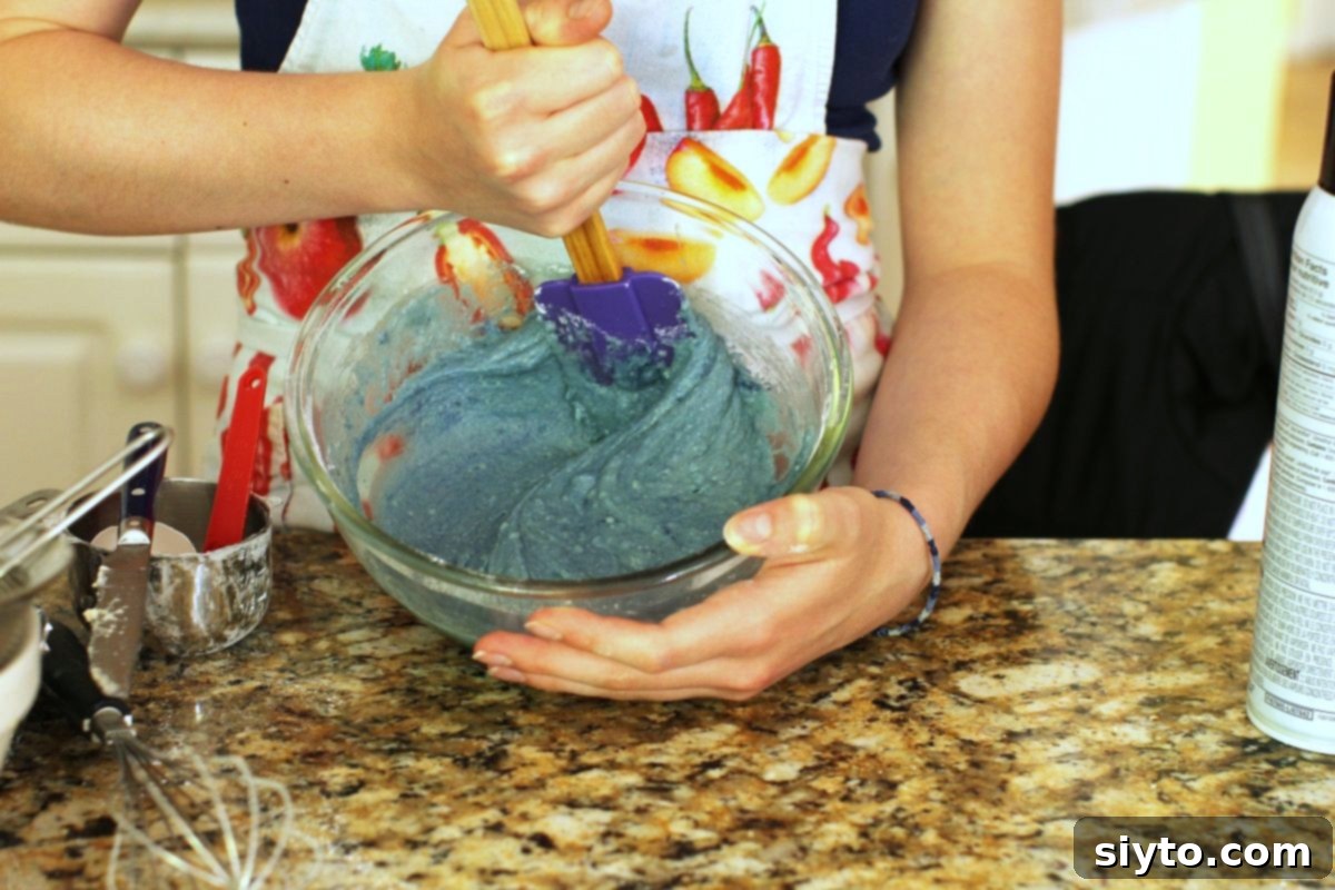 A close-up shot of two hands holding a mixing bowl, actively stirring the vivid blue donut batter with a spatula, showcasing its mesmerizing color.