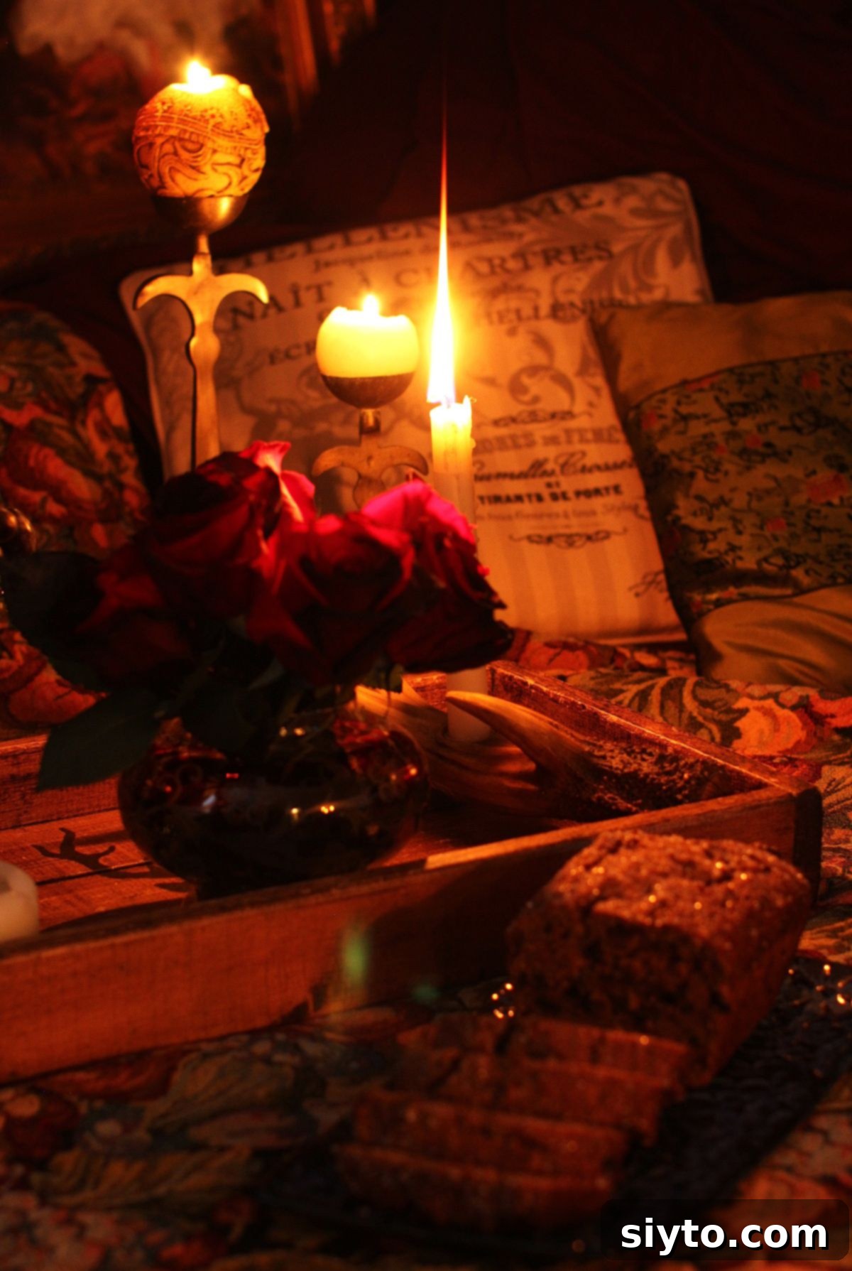 Round candle and short candle glowing in the dark on a wooden tray, with bouquet of dark roses and bara brith cake in front.