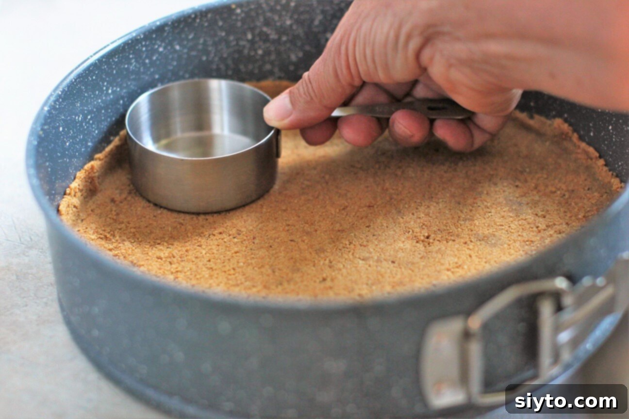 A hand using the bottom of a measuring cup to smoothly and evenly pack down the graham cracker crust into the springform pan.